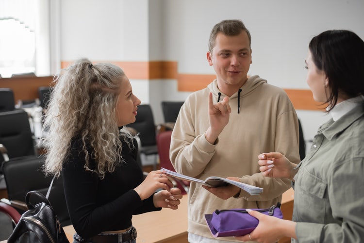 Three young people stand together in a classroom or lecture hall, communicating using sign language while holding notebooks and a purple pencil case.