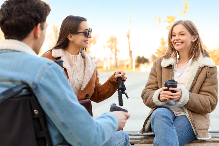 Three friends—including a man in a wheelchair and a woman with a hearing aid—sit outdoors chatting and holding coffee cups on a sunny day.